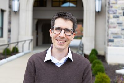 A man wearing glasses and a brown sweater over a collared shirt stands in front of a building, smiling.