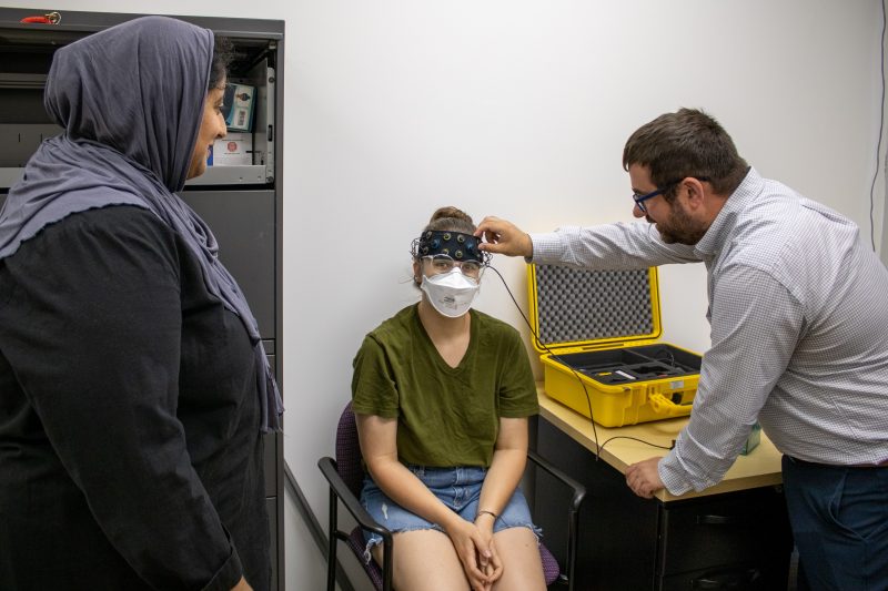 Marrium Mansoor, left, observes Ben Katz, right, as he demonstrates how to use the fNIRS machine, which Diane Devine, center, is wearing on her head.