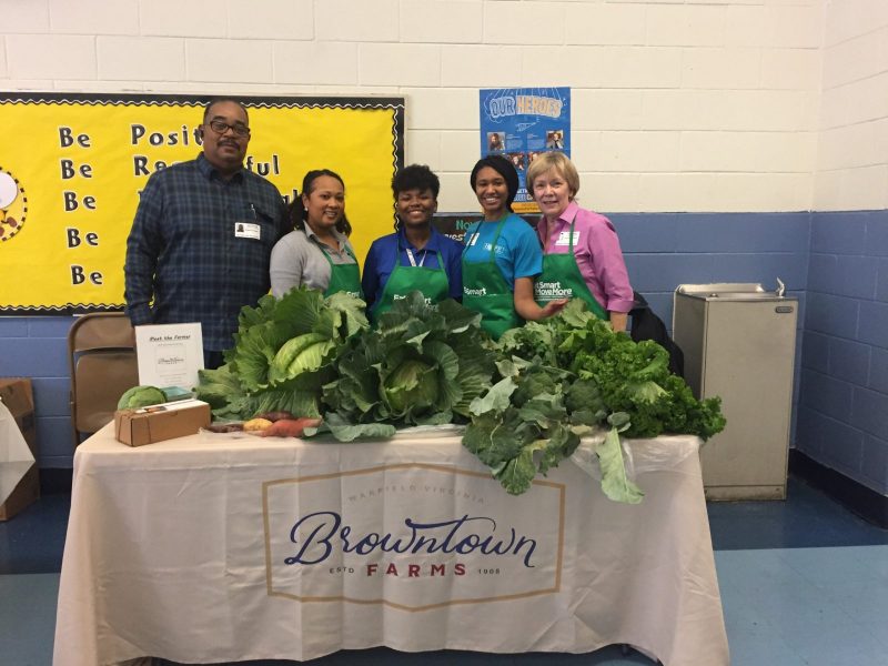 Five people, including four women and one man, posing behind a table piled high with fresh garden produce at caabbage taste testing activity at Pleasants Elementary School in Petersburg, VA.